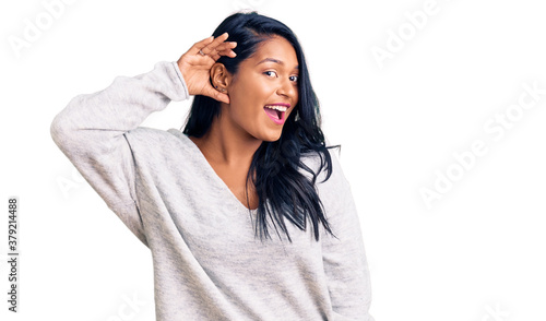 Hispanic woman with long hair wearing casual clothes smiling with hand over ear listening an hearing to rumor or gossip. deafness concept.