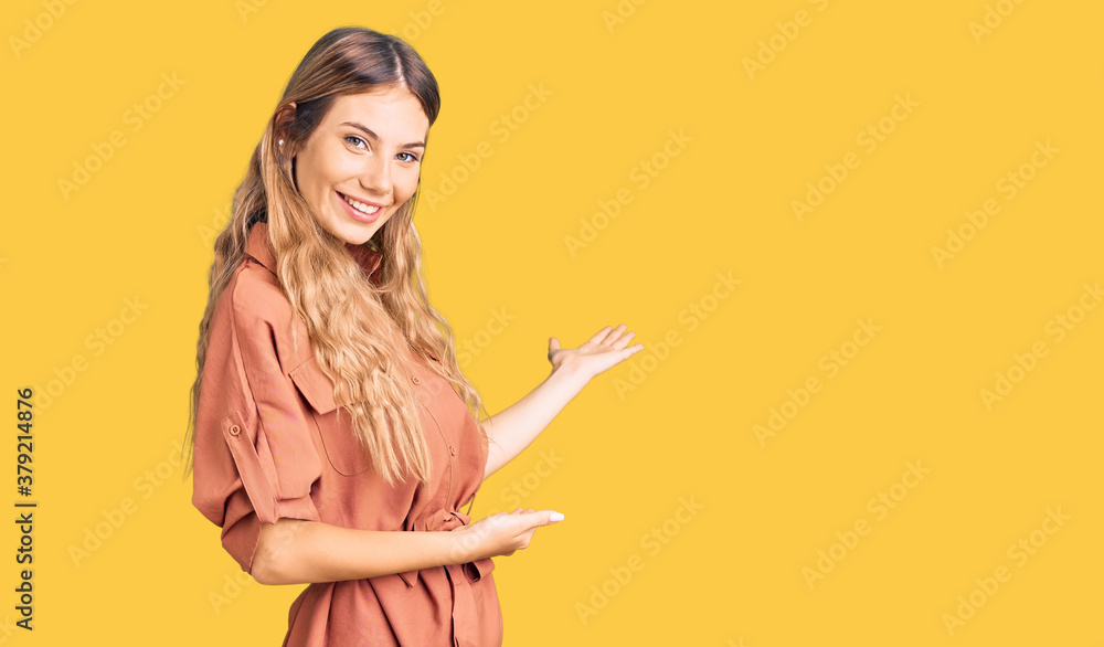 Beautiful caucasian woman with blonde hair wearing summer jumpsuit inviting to enter smiling natural with open hand Beautiful caucasian woman with blonde hair wearing summer jumpsuit inviting to enter smiling natural with open hand