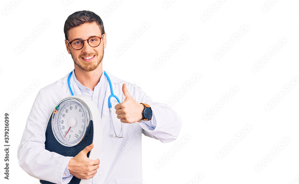 Handsome young man with bear as nutritionist doctor holding weighing machine smiling happy and positive, thumb up doing excellent and approval sign Handsome young man with bear as nutritionist doctor holding weighing machine smiling happy and positive, thumb up doing excellent and approval sign