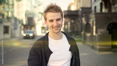 Young smiling guy dressed casually on the background of city road. Middle shot of smiling man standing near the road, on background of blurred residential houses