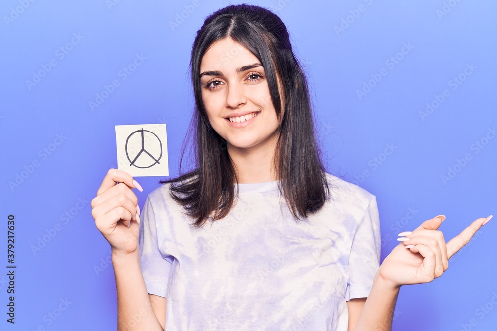 Young beautiful girl holding peace symbol reminder smiling happy pointing with hand and finger to the side Young beautiful girl holding peace symbol reminder smiling happy pointing with hand and finger to the side