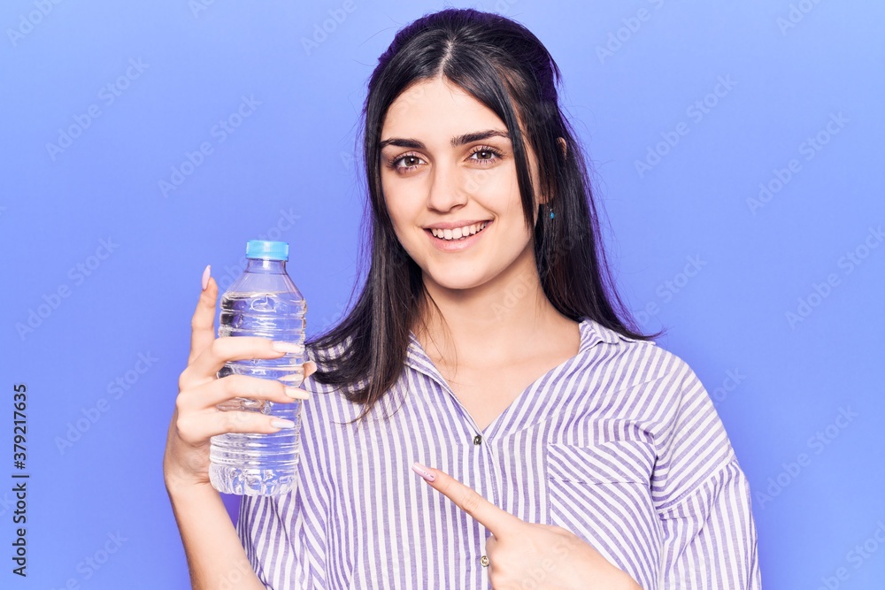Young beautiful girl holding bottle of water smiling happy pointing with hand and finger Young beautiful girl holding bottle of water smiling happy pointing with hand and finger