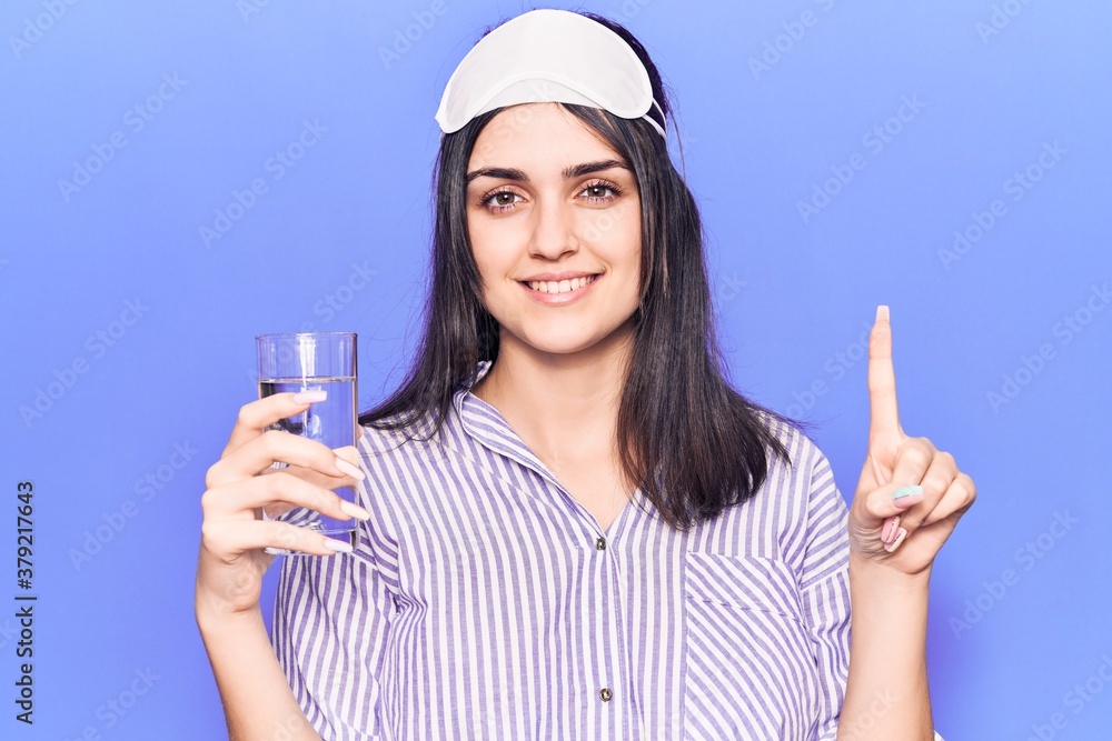 Young beautiful girl wearing sleep mask drinking glass of water smiling with an idea or question pointing finger with happy face, number one Young beautiful girl wearing sleep mask drinking glass of water smiling with an idea or question pointing finger with happy face, number one