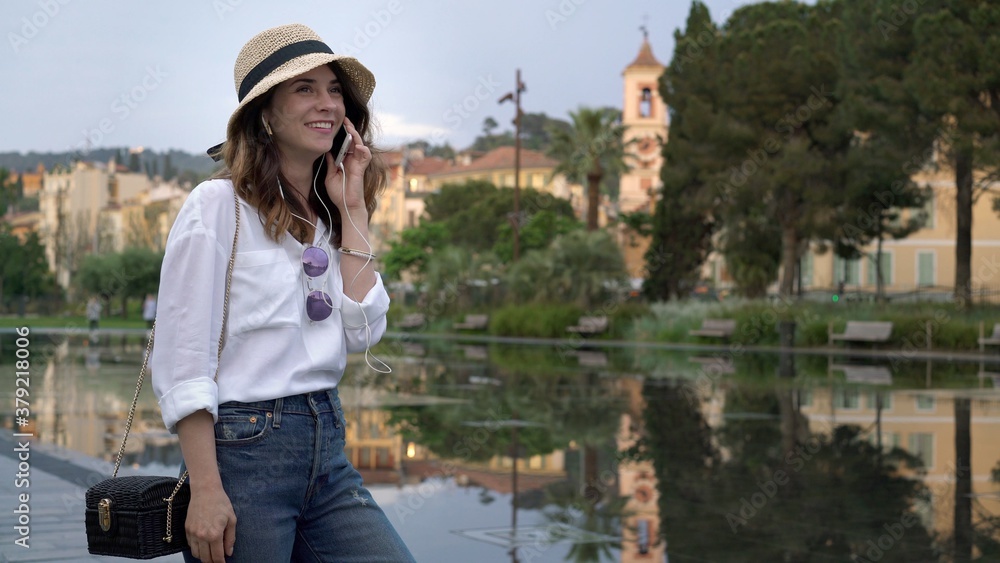 Young woman in hat and white shirt happy talking on a phone in Nice, France. Female tourist in hat and jeans on the phone, on background of green trees and pond