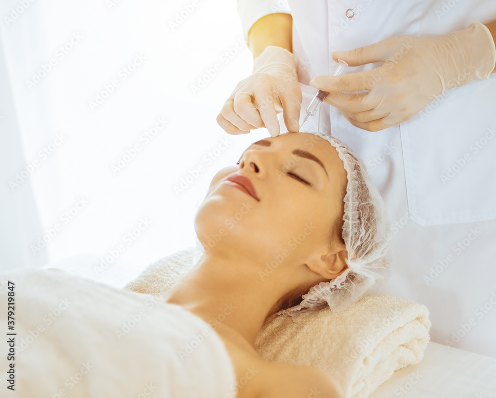 Beautiful woman receiving beauty injections with closed eyes in sunny medical center. Beautician doctor hands doing beauty procedure to female face with syringe