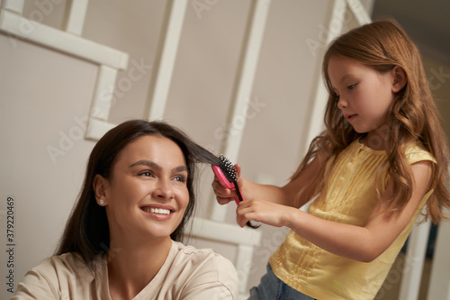 Young woman, happy lovely mother looking aside and smiling while her little daughter combing hair