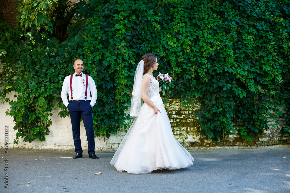 The bride in a white dress in the foreground and the groom with suspenders stands behind. on the background wall with green leaves