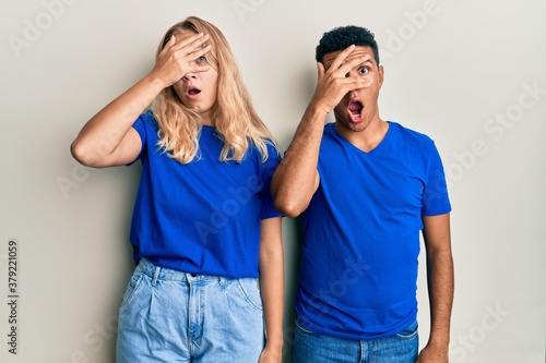 Young interracial couple wearing casual clothes peeking in shock covering face and eyes with hand, looking through fingers with embarrassed expression.