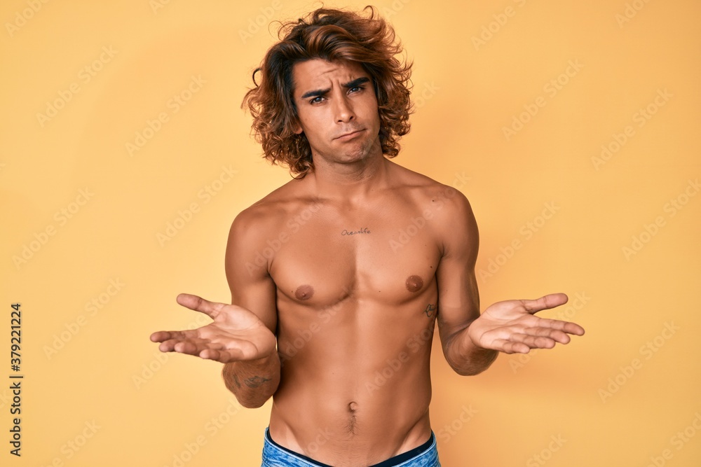 Young hispanic man standing shirtless clueless and confused with open arms, no idea concept.