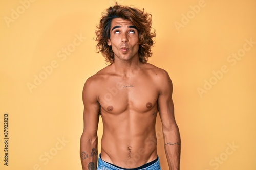 Young hispanic man standing shirtless making fish face with lips  crazy and comical gesture. funny expression.