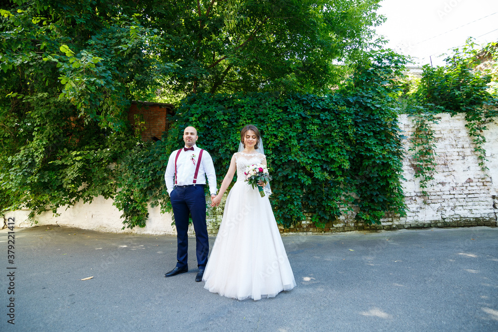 Beautiful couple bride in a white dress with a bouquet while the groom with suspenders and bow tie holding hands. Against the background of a wall with green leaves. Happy couple. Marriage concept. Beautiful couple bride in a white dress with a bouquet while the groom with suspenders and bow tie holding hands. Against the background of a wall with green leaves. Happy couple. Marriage concept.