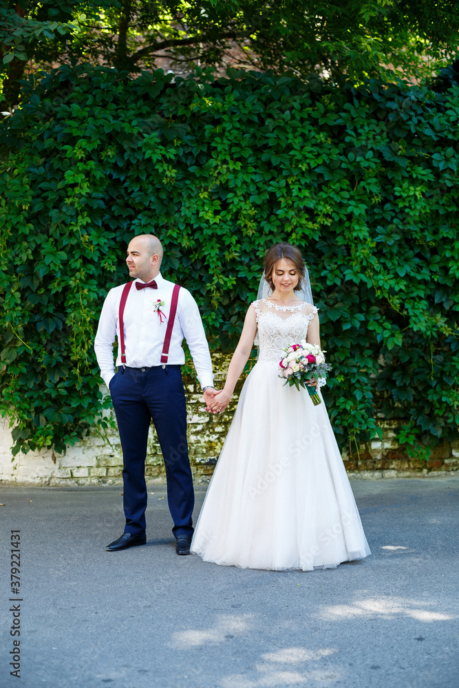 Beautiful couple bride in a white dress with a bouquet while the groom with suspenders and bow tie holding hands. Against the background of a wall with green leaves. Happy couple. Marriage concept. Beautiful couple bride in a white dress with a bouquet while the groom with suspenders and bow tie holding hands. Against the background of a wall with green leaves. Happy couple. Marriage concept.