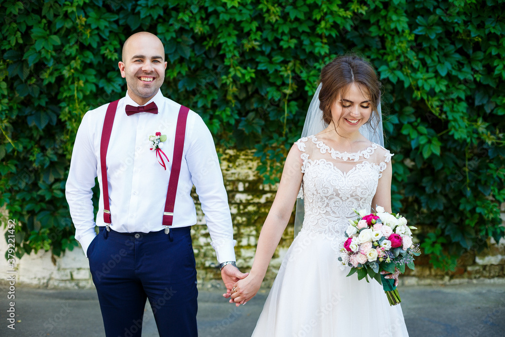 Beautiful couple bride in a white dress with a bouquet while the groom with suspenders and bow tie holding hands. Against the background of a wall with green leaves. Happy couple. Marriage concept.
