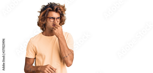 Young hispanic man wearing casual clothes and glasses smelling something stinky and disgusting, intolerable smell, holding breath with fingers on nose. bad smell