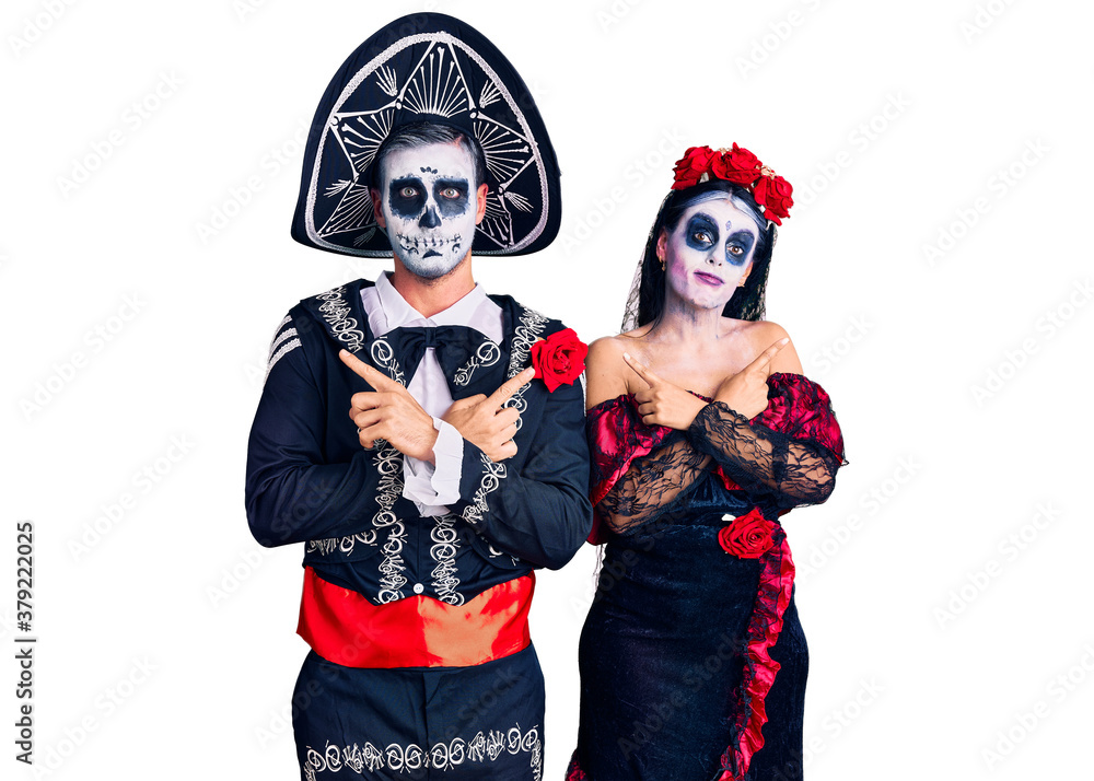 Young couple wearing mexican day of the dead costume over background pointing to both sides with fingers, different direction disagree