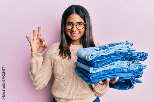 Young latin girl holding stack of folded jeans doing ok sign with fingers  smiling friendly gesturing excellent symbol