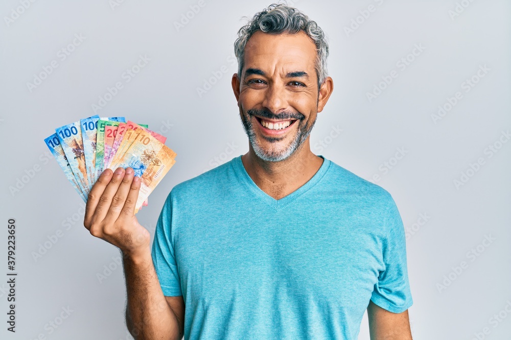 Middle age grey-haired man holding swiss franc banknotes looking positive and happy standing and smiling with a confident smile showing teeth Middle age grey-haired man holding swiss franc banknotes looking positive and happy standing and smiling with a confident smile showing teeth