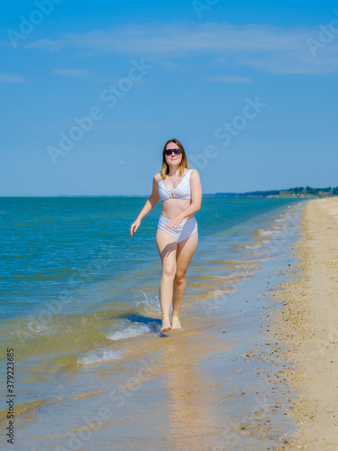 A young girl runs along the sandy sea beach along the surf. Splashes of sea water. The girl is wearing a white swimsuit and sunglasses. Freedom and carelessness. Outdoor fitness. Sunny day. Copy space