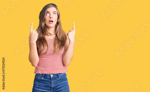 Beautiful caucasian young woman wearing casual clothes amazed and surprised looking up and pointing with fingers and raised arms.