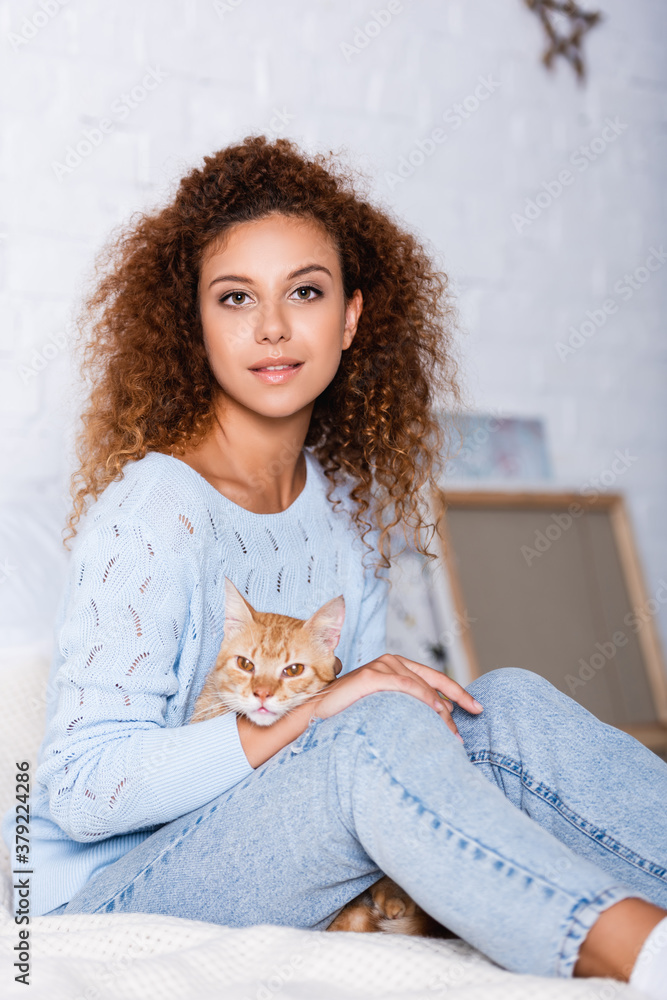Selective focus of young curly woman looking at camera while holding cat on bed Selective focus of young curly woman looking at camera while holding cat on bed