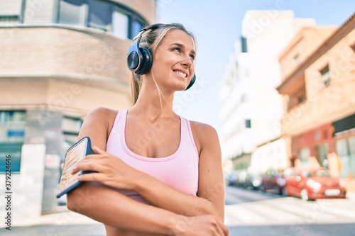 Young blonde sporty girl using headphones walking at street of city.