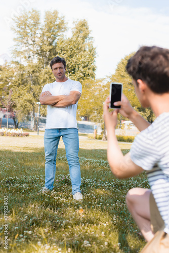 selective focus of man standing with crossed arms while teenager boy taking photo and holding smartphone in park