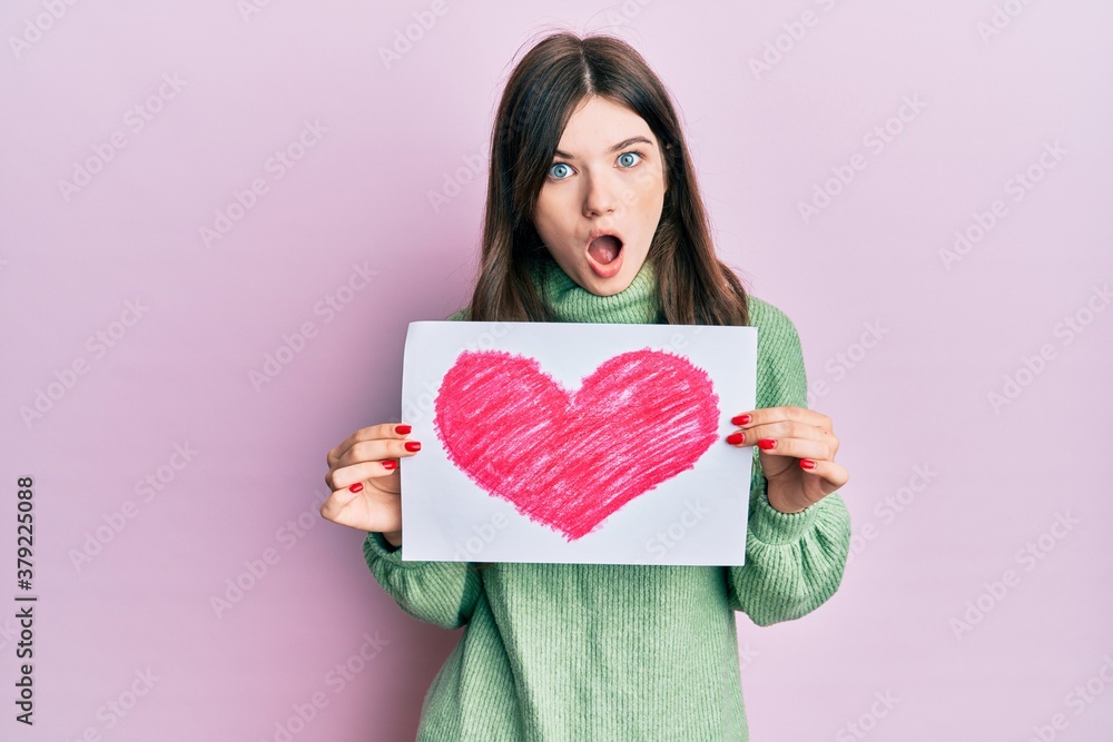 Young beautiful caucasian girl holding heart draw afraid and shocked with surprise and amazed expression, fear and excited face. Young beautiful caucasian girl holding heart draw afraid and shocked with surprise and amazed expression, fear and excited face.