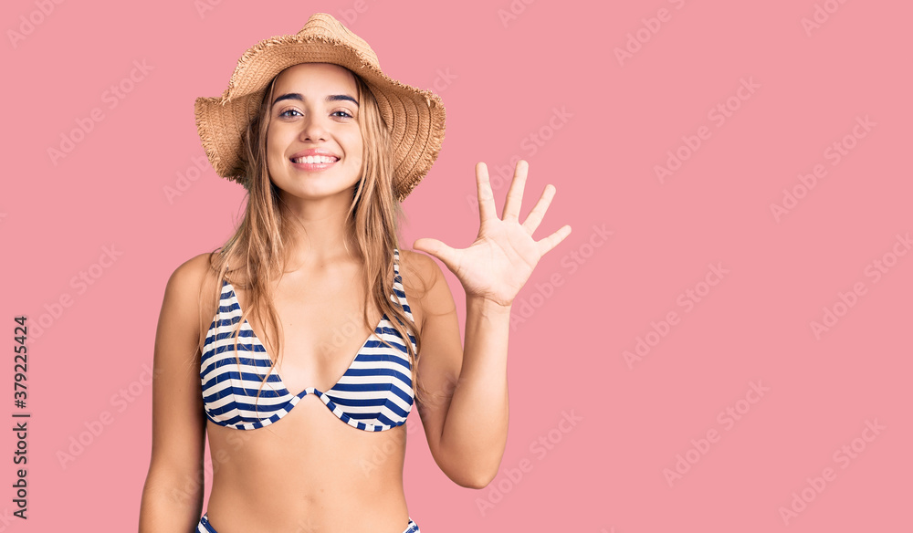 Naklejka premium Young beautiful blonde woman wearing bikini and hat showing and pointing up with fingers number five while smiling confident and happy.