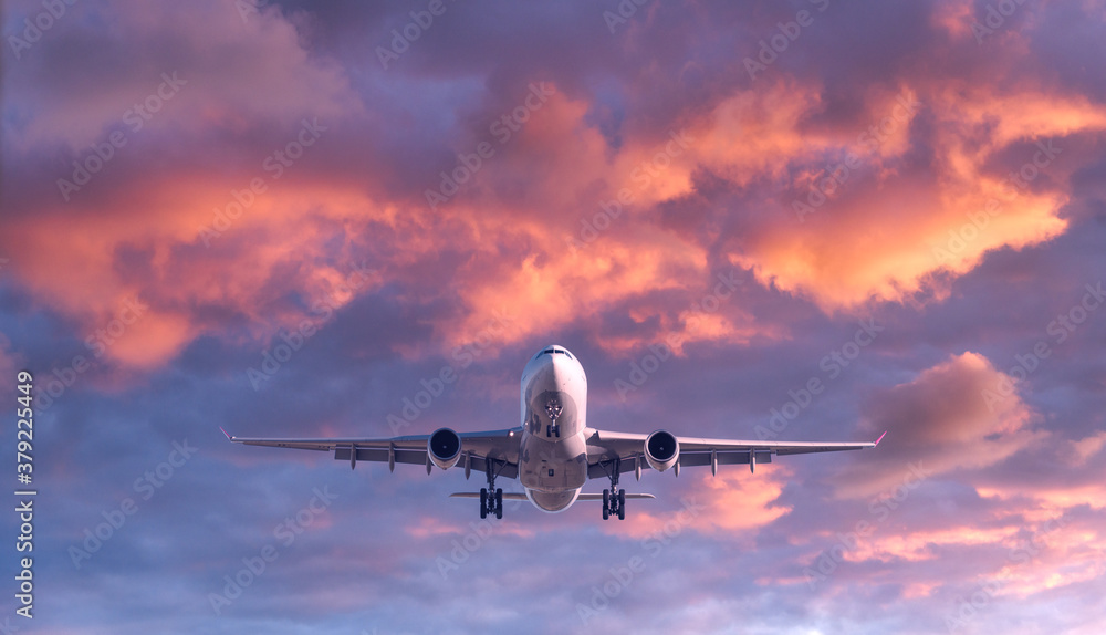 Airplane is flying in colorful sky at sunset. Landscape with white passenger airplane, purple sky with pink clouds. Aircraft is landing. Business trip. Commercial plane. Travel. Aerial view. Concept