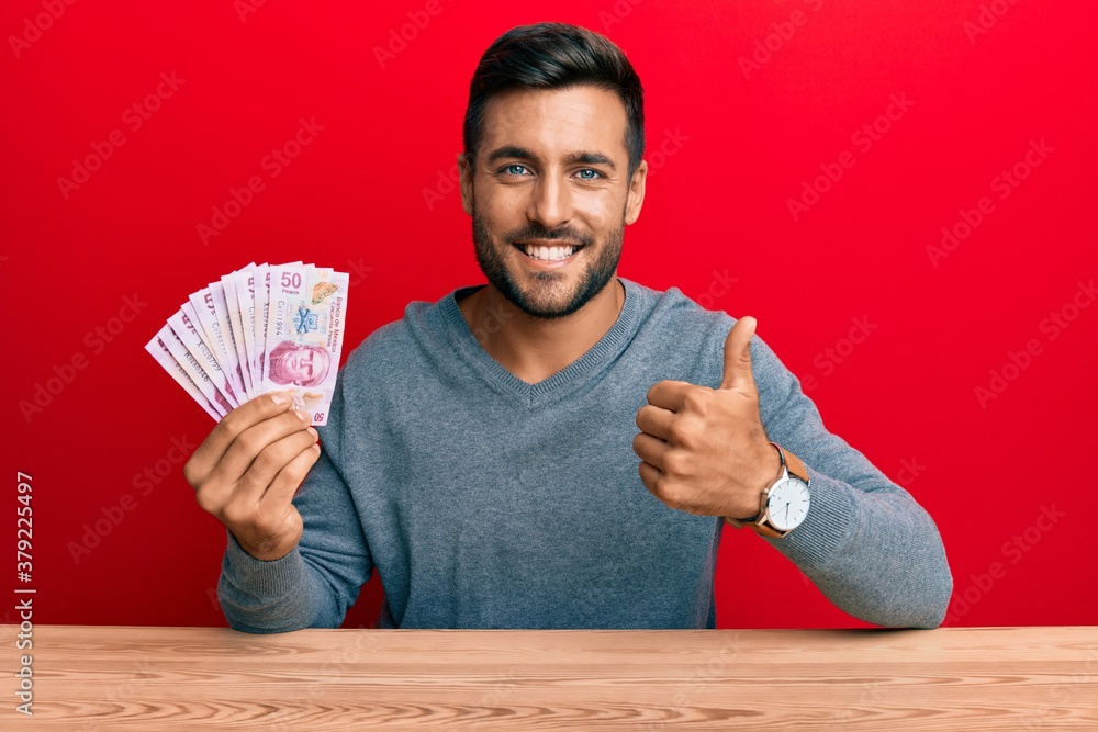 Handsome hispanic man holding mexican pesos smiling happy and positive, thumb up doing excellent and approval sign Handsome hispanic man holding mexican pesos smiling happy and positive, thumb up doing excellent and approval sign