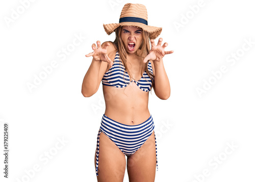 Young beautiful blonde woman wearing bikini and hat smiling funny doing claw gesture as cat, aggressive and sexy expression