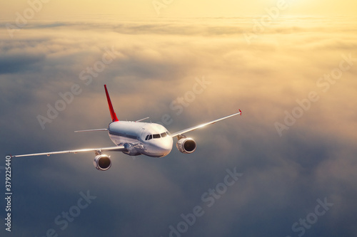 Airplane is flying above the clouds at sunset in summer. Landscape with passenger airplane, low clouds, orange sky. Front view of aircraft. Business travel. Commercial plane. Aerial view. Take off