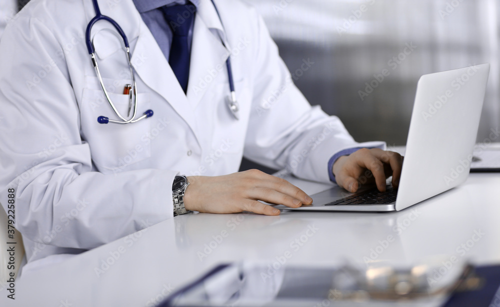 Unknown male doctor sitting and working with laptop in clinic at his working place, close-up. Young physician at work. Perfect medical service, medicine concept Unknown male doctor sitting and working with laptop in clinic at his working place, close-up. Young physician at work. Perfect medical service, medicine concept