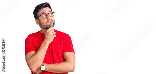 Young hispanic man wearing casual clothes and glasses serious face thinking about question with hand on chin, thoughtful about confusing idea