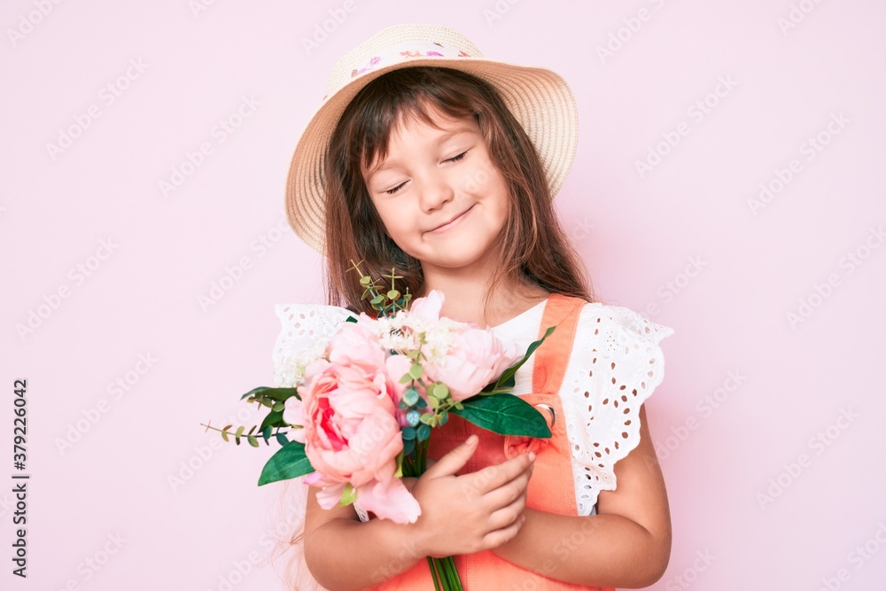 Little caucasian kid girl with long hair holding flower wearing spring hat looking positive and happy standing and smiling with a confident smile showing teeth Little caucasian kid girl with long hair holding flower wearing spring hat looking positive and happy standing and smiling with a confident smile showing teeth