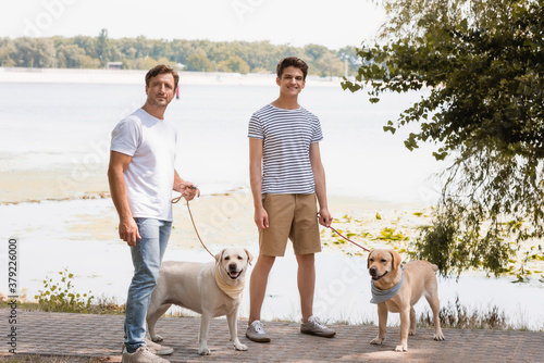 father and teenager son holding leashes and standing with golden retrievers near lake