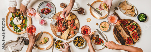 Friends wine and snacks party. Flay-lay of peoples hands with rose wine and food over table with cheese, fruit, smoked meat, tomato brushettas, buratta salad, top view. Wine tasting, gathering concept