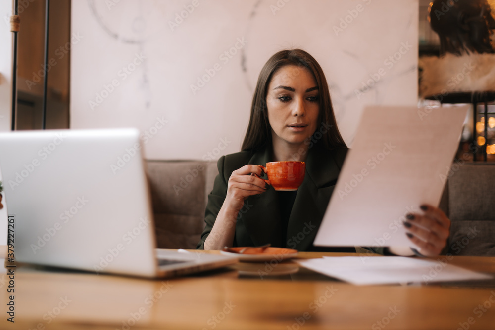 Pretty young business woman studying paper documents in cafe sitting at table with laptop. Lady enjoys delicious coffee while working. Student girl sitting in cafe with cup of coffee. Pretty young business woman studying paper documents in cafe sitting at table with laptop. Lady enjoys delicious coffee while working. Student girl sitting in cafe with cup of coffee.