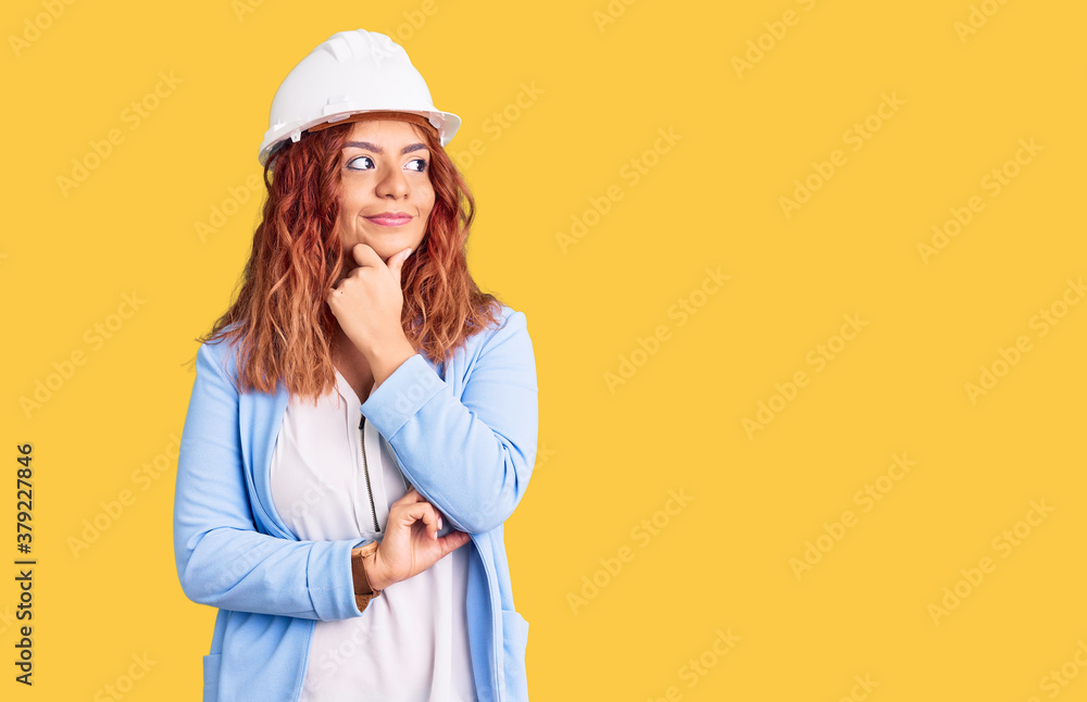 Young latin woman wearing architect hardhat with hand on chin thinking about question, pensive expression. smiling with thoughtful face. doubt concept. Young latin woman wearing architect hardhat with hand on chin thinking about question, pensive expression. smiling with thoughtful face. doubt concept.