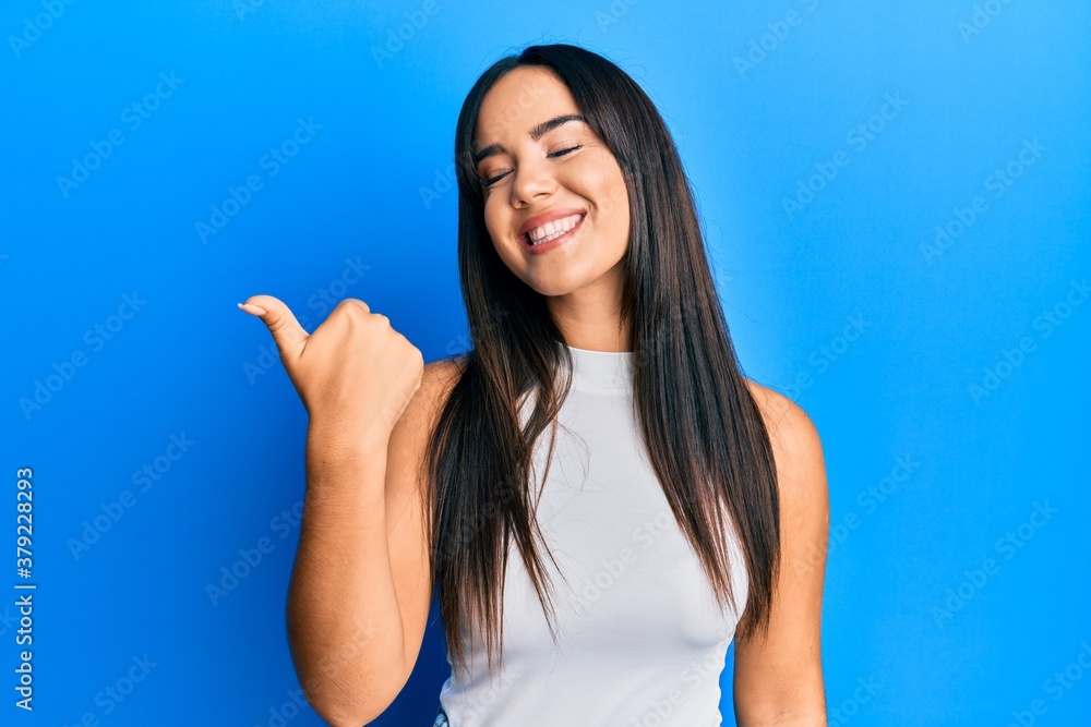 Young beautiful hispanic girl wearing casual clothes pointing thumb up to the side smiling happy with open mouth Young beautiful hispanic girl wearing casual clothes pointing thumb up to the side smiling happy with open mouth