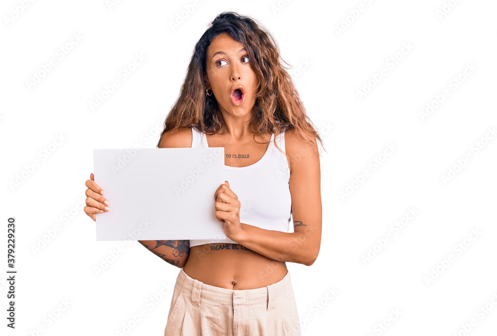 Young hispanic woman with tattoo holding blank empty paper scared and amazed with open mouth for surprise, disbelief face Young hispanic woman with tattoo holding blank empty paper scared and amazed with open mouth for surprise, disbelief face