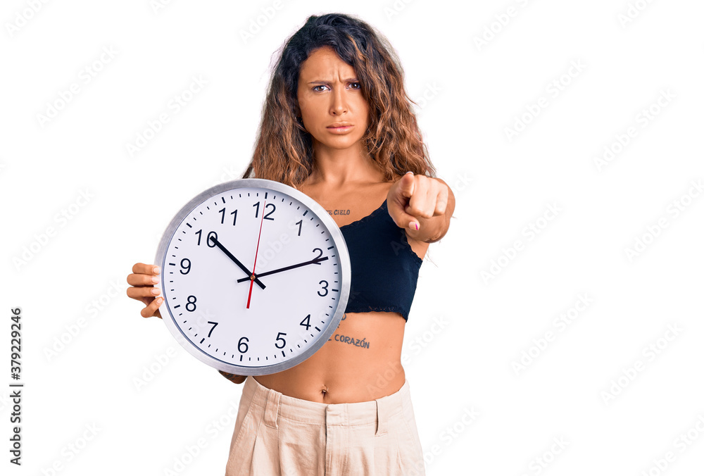 Young hispanic woman with tattoo holding big clock pointing with finger to the camera and to you, confident gesture looking serious Young hispanic woman with tattoo holding big clock pointing with finger to the camera and to you, confident gesture looking serious
