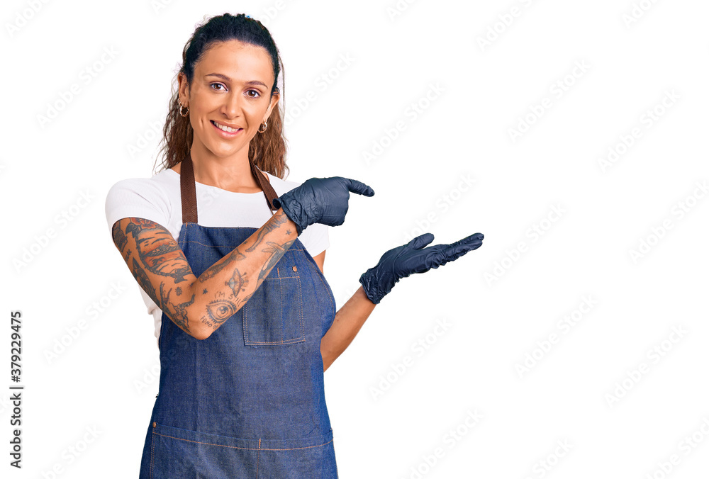 Young hispanic woman with tattoo wearing barber apron and gloves amazed and smiling to the camera while presenting with hand and pointing with finger. Young hispanic woman with tattoo wearing barber apron and gloves amazed and smiling to the camera while presenting with hand and pointing with finger.