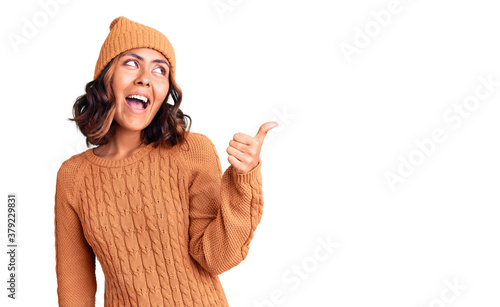 Young beautiful mixed race woman wearing wool sweater and winter hat smiling with happy face looking and pointing to the side with thumb up. photo