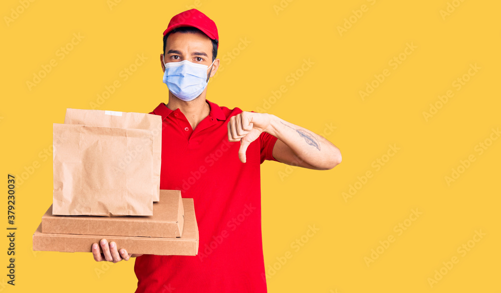 Young handsome hispanic man delivering food wearing covid-19 safety mask with angry face, negative sign showing dislike with thumbs down, rejection concept Young handsome hispanic man delivering food wearing covid-19 safety mask with angry face, negative sign showing dislike with thumbs down, rejection concept