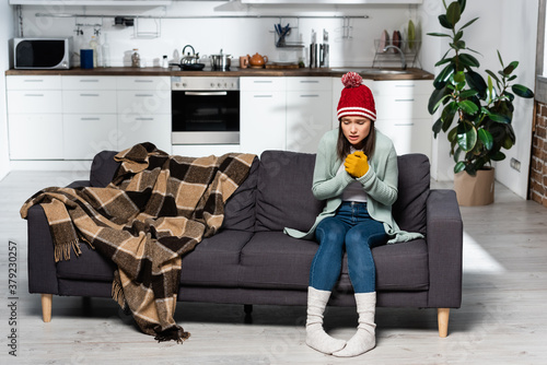 Photography shivering woman in warm hat, gloves and socks sitting on sofa near plaid blanket
