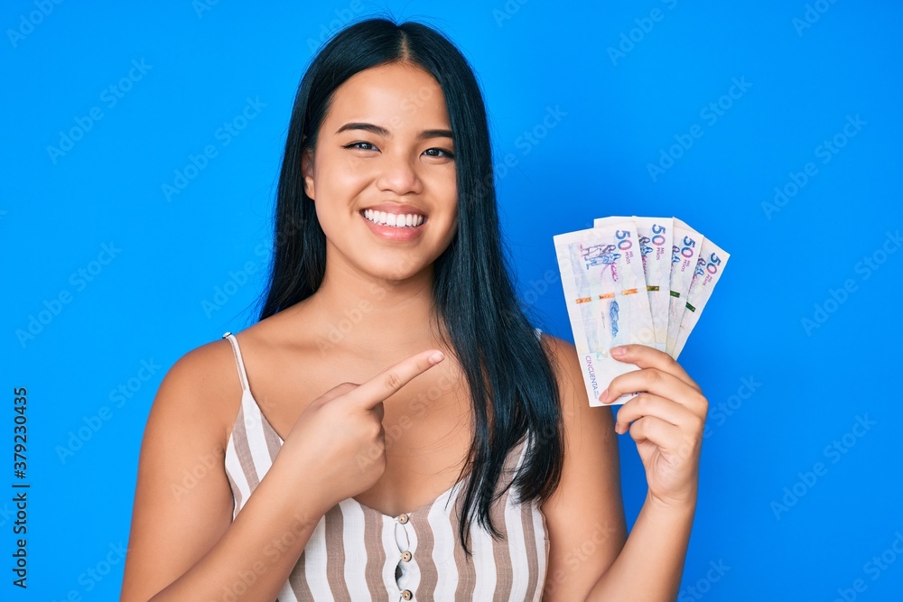Young beautiful asian girl holding colombian pesos smiling happy pointing with hand and finger Young beautiful asian girl holding colombian pesos smiling happy pointing with hand and finger
