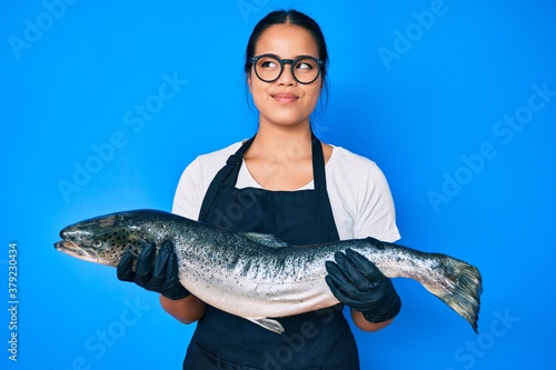 Young beautiful asian girl fishmonger selling fresh raw salmon smiling looking to the side and staring away thinking.