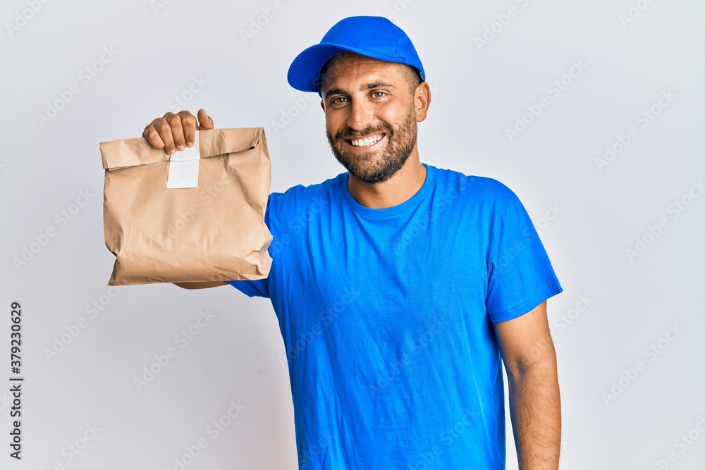Handsome man with beard holding delivery package looking positive and happy standing and smiling with a confident smile showing teeth Handsome man with beard holding delivery package looking positive and happy standing and smiling with a confident smile showing teeth