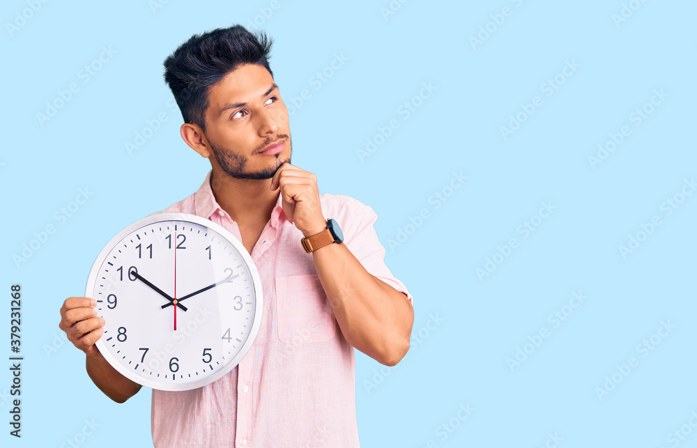 Handsome latin american young man holding big clock serious face thinking about question with hand on chin, thoughtful about confusing idea Handsome latin american young man holding big clock serious face thinking about question with hand on chin, thoughtful about confusing idea